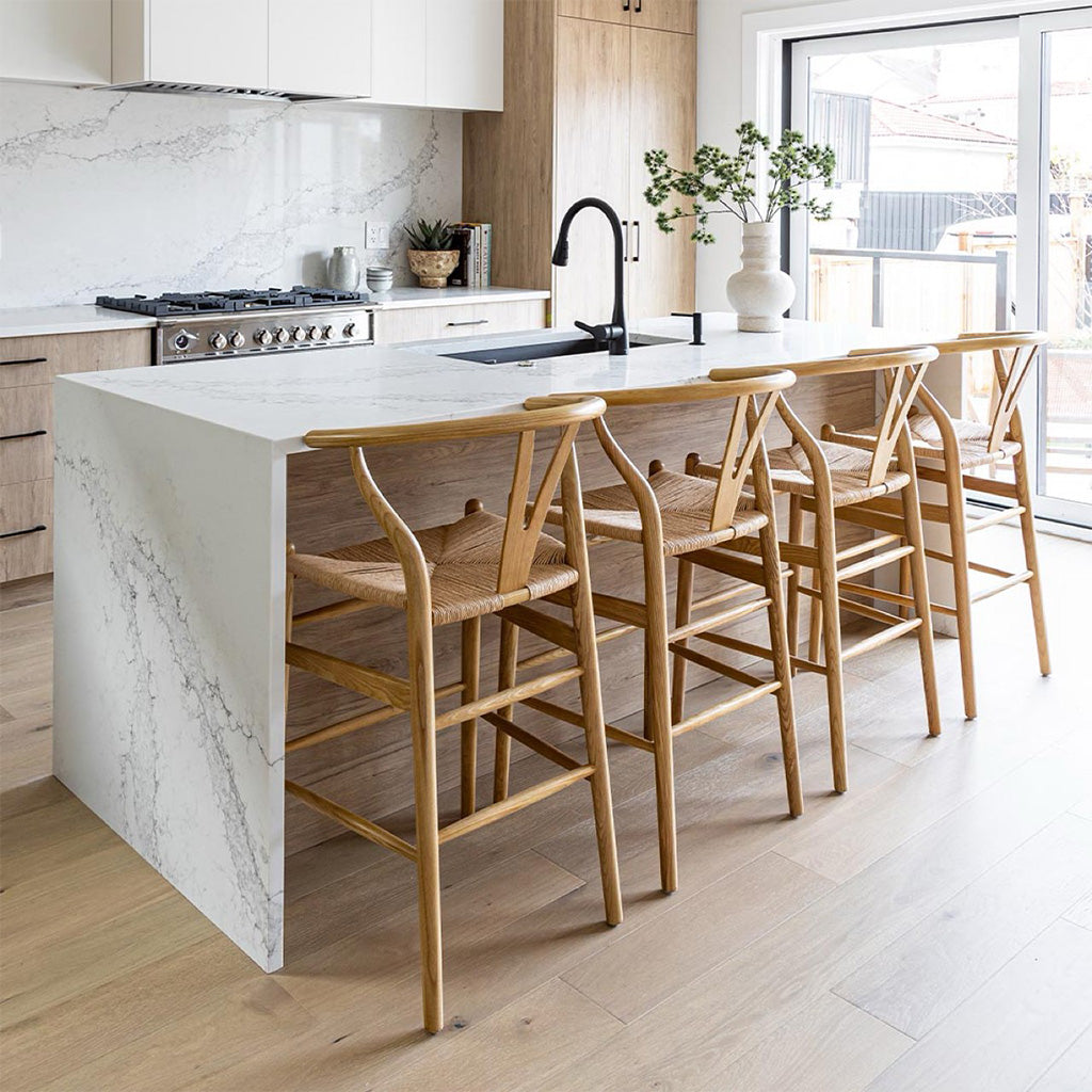 4 Frida Counter Stools in blonde shown at marble island. A minimalist counter stool inspired by the mid-mod 1950s Hans J Wegner wishbone chairs with a Y-back design, curving top rail and tapered legs details.